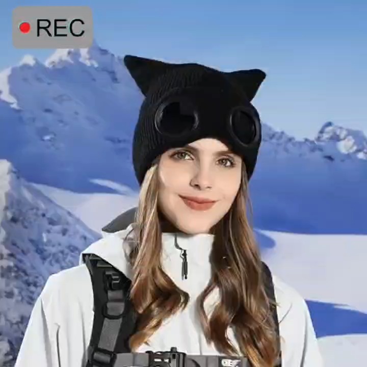 Woman wearing a black cat ear knit hat with round dark lenses against a snowy mountain backdrop.